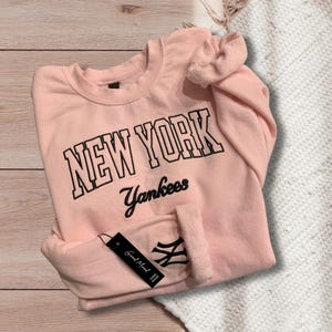 May include: A pink sweatshirt with the text "NEW YORK Yankees" in black. The sweatshirt has long sleeves and a crew neck.