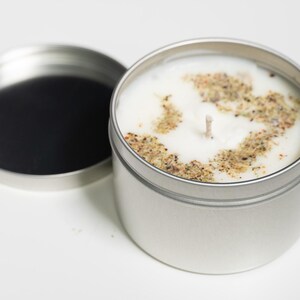 以下が含まれることがあります： 銀色のキャンドル缶に白いキャンドルが入っています。キャンドルの上には乾燥ハーブの層があります。