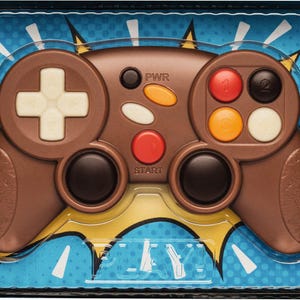 Puede incluir: Un mando de videojuego de chocolate marrón con botones coloridos y una cruceta blanca en forma de cruz. El mando está sobre un fondo azul con detalles de cómic. La palabra "PLAY" es visible.
