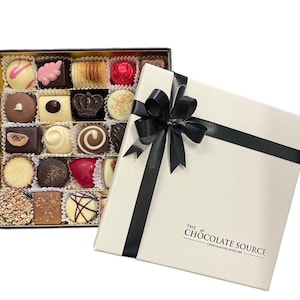 May include: A white box with a black ribbon tied in a bow. The box is labelled "The Chocolate Source" and contains a selection of chocolates in a black and gold box.