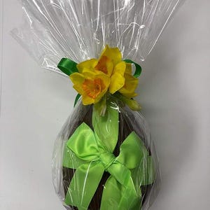 Può includere: Un uovo di cioccolato fondente avvolto in cellophane trasparente, adornato con un fiocco di raso verde brillante e un grappolo di narcisi gialli. Un regalo festivo per Pasqua o un'occasione speciale.