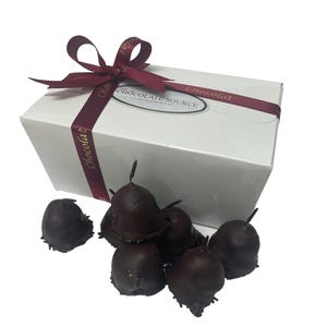 May include: A white box with a red ribbon tied around it. The box is labeled "The Chocolate Source" and has a red ribbon with the word "Chocolat" printed on it. The box is surrounded by a pile of dark chocolate covered cherries.