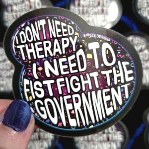 May include: A black sticker with a white border and the text "I DON'T NEED THERAPY I NEED TO FIST FIGHT THE GOVERNMENT" in white. The background features purple, pink, and yellow confetti.