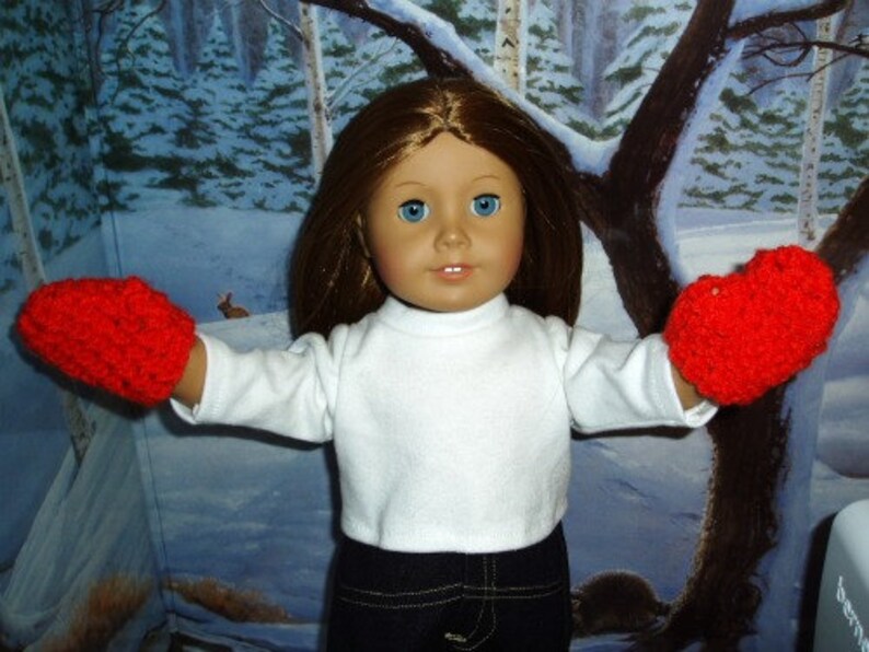 K&ouml;nnte beinhalten: Eine Puppe tr&auml;gt ein wei&szlig;es Langarmshirt und rote H&auml;kelhandschuhe. Die Puppe hat langes braunes Haar und blaue Augen. Die Puppe steht vor einem verschneiten Hintergrund.