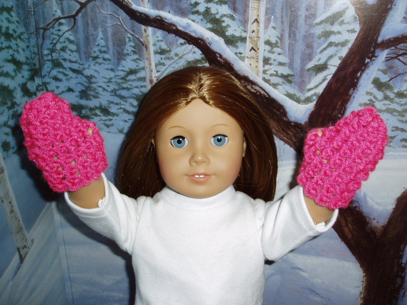 K&ouml;nnte beinhalten: Eine Puppe tr&auml;gt ein wei&szlig;es Langarmhemd und rosa geh&auml;kelte F&auml;ustlinge. Die Puppe hat langes braunes Haar und blaue Augen.