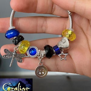 Puede incluir: Una pulsera de plata con abalorios que presenta varias cuentas y dijes. La pulsera incluye cuentas azules, amarillas y negras, un dije de lib&eacute;lula, un dije de bot&oacute;n y un dije de estrella. La imagen tambi&eacute;n incluye un cartel de la pel&iacute;cula "Coraline."