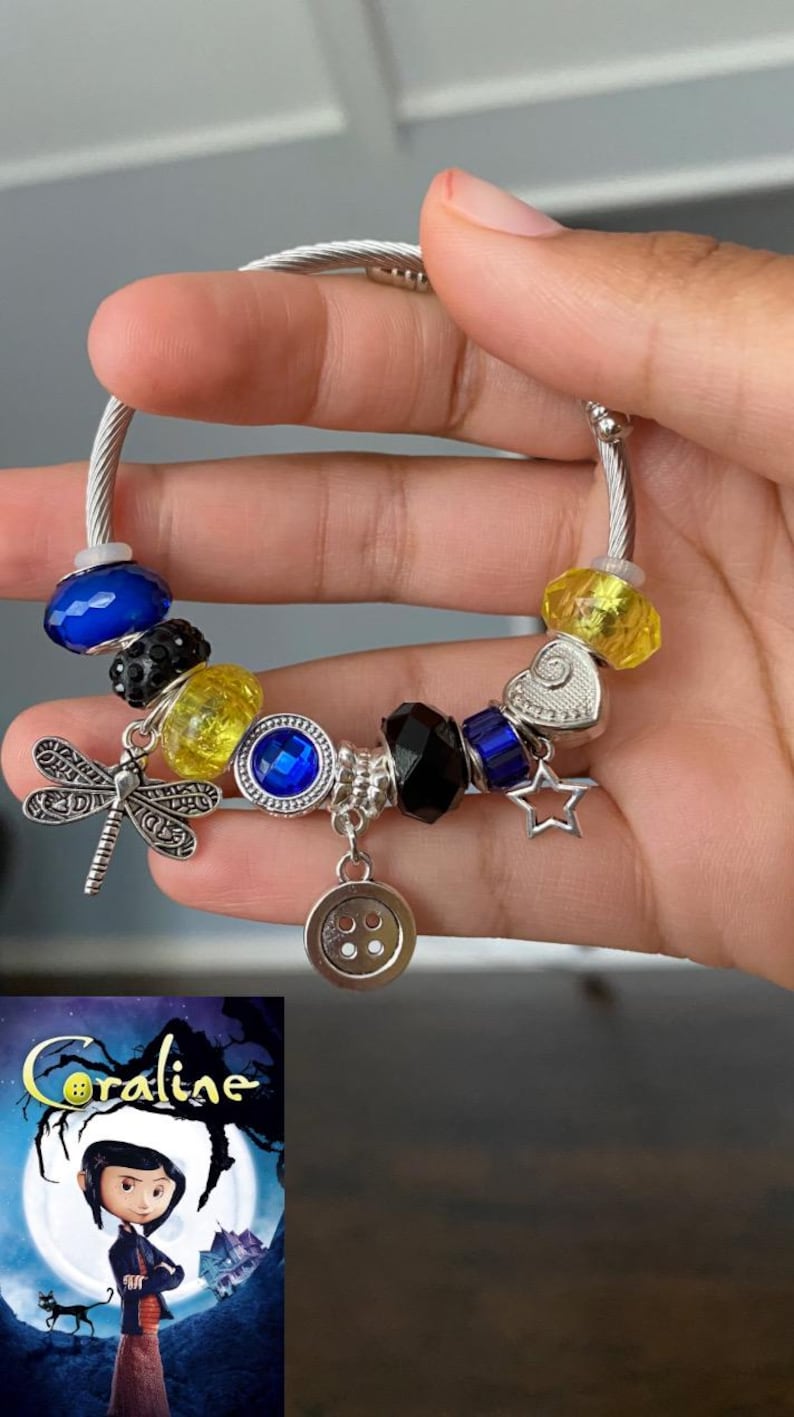 Puede incluir: Una pulsera de plata con abalorios que presenta varias cuentas y dijes. La pulsera incluye cuentas azules, amarillas y negras, un dije de lib&eacute;lula, un dije de bot&oacute;n y un dije de estrella. La imagen tambi&eacute;n incluye un cartel de la pel&iacute;cula "Coraline."
