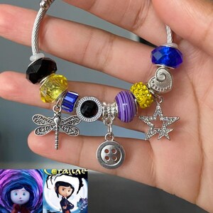 Puede incluir: Pulsera de abalorios plateada con varias cuentas y dijes. Incluye una lib&eacute;lula, un bot&oacute;n, una estrella y un dije de coraz&oacute;n. Las cuentas son de color negro, amarillo, azul y morado. La pulsera se sostiene en una mano, con im&aacute;genes de la pel&iacute;cula "Coraline" en el fondo.