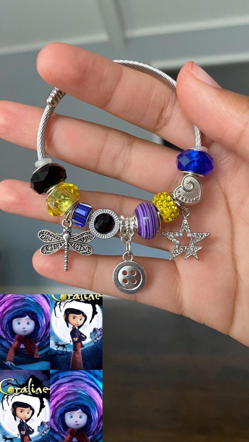 Puede incluir: Pulsera de abalorios plateada con varias cuentas y dijes. Incluye una lib&eacute;lula, un bot&oacute;n, una estrella y un dije de coraz&oacute;n. Las cuentas son de color negro, amarillo, azul y morado. La pulsera se sostiene en una mano, con im&aacute;genes de la pel&iacute;cula "Coraline" en el fondo.