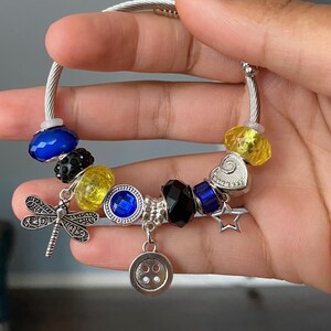 Puede incluir: Pulsera de abalorios plateada con varias cuentas y abalorios de colores. Incluye cuentas azules, amarillas y negras, un abalorio de lib&eacute;lula, un abalorio de carita sonriente y un abalorio de estrella. La pulsera se sostiene en una mano.