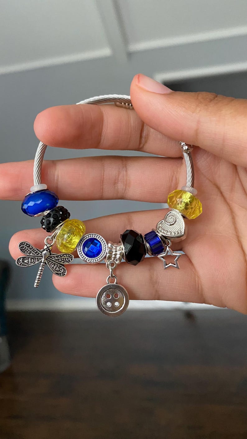 Puede incluir: Pulsera de abalorios plateada con varias cuentas y abalorios de colores. Incluye cuentas azules, amarillas y negras, un abalorio de lib&eacute;lula, un abalorio de carita sonriente y un abalorio de estrella. La pulsera se sostiene en una mano.