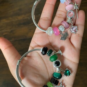 May include: Two silver-tone charm bracelets, each adorned with colorful beads and charms. One bracelet features pink and clear beads, a crown, and a heart charm. The other has green and black beads, a star charm, and a green gem.