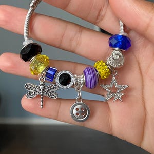 Puede incluir: Una pulsera de plata con varios abalorios y cuentas de colores. La pulsera presenta un dije de lib&eacute;lula, un dije de bot&oacute;n, un dije de estrella y cuentas facetadas en azul, amarillo, negro y morado. La pulsera se sostiene en una mano.