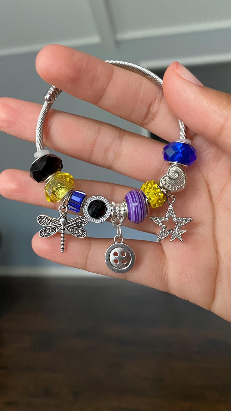 Puede incluir: Una pulsera de plata con varios abalorios y cuentas de colores. La pulsera presenta un dije de lib&eacute;lula, un dije de bot&oacute;n, un dije de estrella y cuentas facetadas en azul, amarillo, negro y morado. La pulsera se sostiene en una mano.
