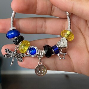 Puede incluir: Pulsera de abalorios plateada con varias cuentas y abalorios. La pulsera presenta cuentas azules, amarillas y negras, junto con un abalorio de lib&eacute;lula, un abalorio de bot&oacute;n y un abalorio de estrella. La pulsera se sostiene en una mano.