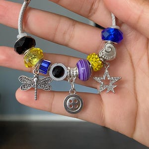 Puede incluir: Una pulsera plateada con una variedad de abalorios coloridos. Incluye una lib&eacute;lula, una estrella, un bot&oacute;n y cuentas negras, amarillas, azules y moradas. La pulsera se sostiene en una mano.