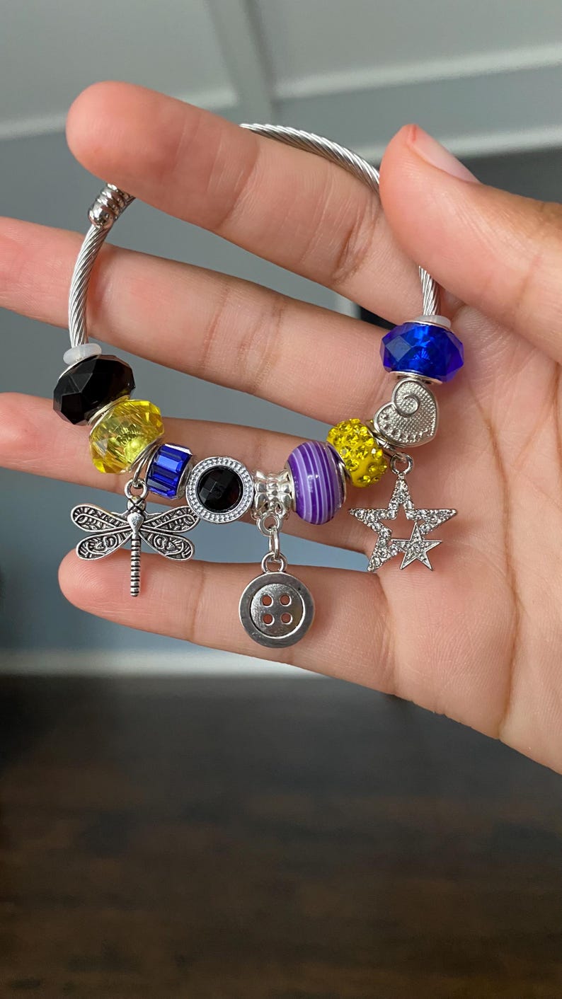 Puede incluir: Una pulsera plateada con una variedad de abalorios coloridos. Incluye una lib&eacute;lula, una estrella, un bot&oacute;n y cuentas negras, amarillas, azules y moradas. La pulsera se sostiene en una mano.