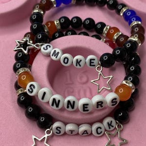 Könnte beinhalten: Drei Armbänder mit Perlen in Schwarz, Braun, Orange und Blau. Eines buchstabiert "SMOKE", ein anderes "SINNERS" und das dritte "STACK". Jedes hat einen silbernen Sternanhänger. Die Armbänder werden auf einer rosa Oberfläche präsentiert.