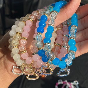 May include: A set of four beaded bracelets with charms. The bracelets are made with clear, pink, and blue beads. The charms are gold-toned and shaped like a cat.