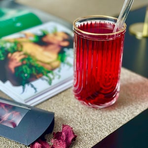 May include: A glass of red juice with a glass straw in a clear glass with a gold rim. The glass is on a brown placemat with a black surface in the background. There are dried beet chips on the placemat.