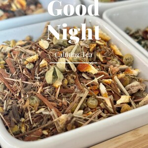 May include: Close-up of a white ceramic bowl filled with a blend of dried herbs, flowers, and fruit peels. The bowl is labeled "Good Night Calming Tea" and is sitting on a wooden surface.