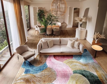 May include: A colorful area rug with an abstract design dominates the room. The rug features flowing shapes in shades of blue, pink, green, and brown. A white sofa with cream-colored pillows sits above the rug. A wooden chair and side table are also visible.