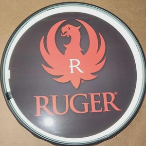 May include: A circular wall sign featuring a dark background with a prominent orange phoenix logo and the letter R centered above the text RUGER in orange capital letters. The sign has a thin white border and a dark outer frame, creating a clean look.
