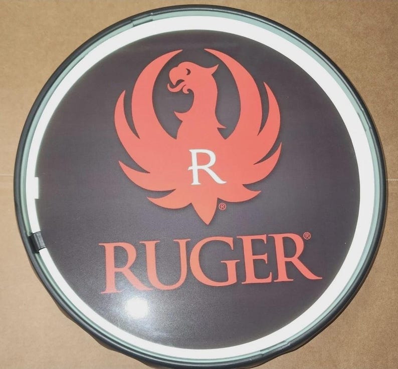May include: A circular wall sign featuring a dark background with a prominent orange phoenix logo and the letter R centered above the text RUGER in orange capital letters. The sign has a thin white border and a dark outer frame, creating a clean look.