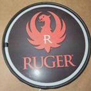 May include: A circular wall sign featuring a black background with a red phoenix logo and the white letter R centered above the red text RUGER. The sign is framed by a thin black border and a white circular light strip along the inner edge.