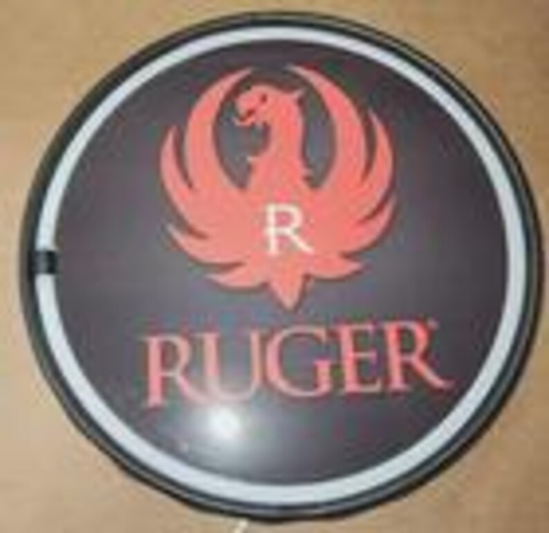 May include: A circular wall sign featuring a black background with a red phoenix logo and the white letter R centered above the red text RUGER. The sign is framed by a thin black border and a white circular light strip along the inner edge.