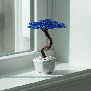 May include: A handcrafted bonsai tree sculpture. The tree features a twisted copper wire trunk and a vibrant blue wire canopy, set in a small, white, crackle-glazed ceramic pot filled with white stones. The bonsai is displayed on a white windowsill.