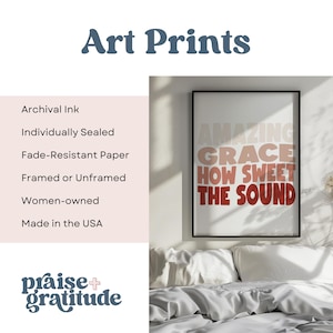 May include: A framed art print with the text "AMAZING GRACE HOW SWEET THE SOUND" in a gradient of pink, orange, and red. The print is on a white background and is framed in a black frame. The print is hanging on a wall above a bed with white bedding.