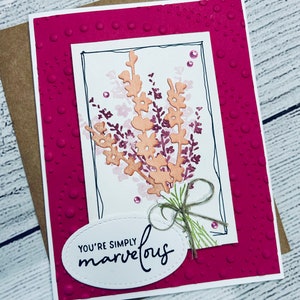 May include: A handmade greeting card with a floral design. The card features a white rectangle with a black border, showcasing peach-colored flowers and dark purple accents. The card reads "YOU'RE SIMPLY marvelous" on an oval label, with a twine bow.
