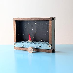 May include: A wooden box diorama featuring a red sailboat on blue waves under a starry night sky. The box has a crank on the side, and the scene is set within a dark interior with a constellation pattern.