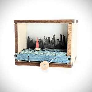 May include: A wooden shadow box featuring a cityscape silhouette with a red-sailed sailboat on blue waves. The box has a light wood frame and a small wooden knob on the side. The background shows a black and white skyline.
