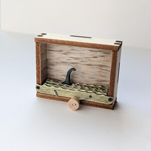 May include: A wooden box with a small, black, wooden figure of a sea monster's head sticking out of the top. The box has a crank on the bottom and a green, wavy, wooden surface inside.