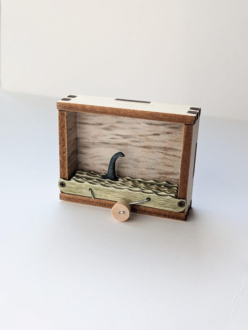 May include: A wooden box with a small, black, wooden figure of a sea monster's head sticking out of the top. The box has a crank on the bottom and a green, wavy, wooden surface inside.