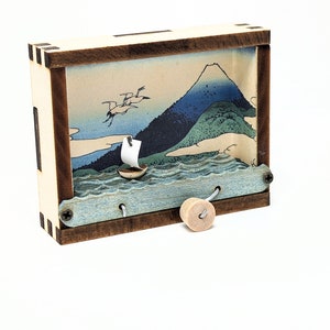 May include: A wooden diorama depicting a sailboat on the water with a mountain backdrop. The diorama is in a rectangular box with a light wood frame and a dark wood trim. A small wooden spool is attached to the diorama.