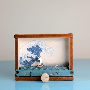 Pode incluir: Caixa de madeira com uma representação da Grande Onda de Kanagawa. A obra de arte utiliza tons de azul e branco, com um pequeno barco na água. A caixa tem uma moldura de madeira e uma base azul em forma de onda com um botão de madeira.