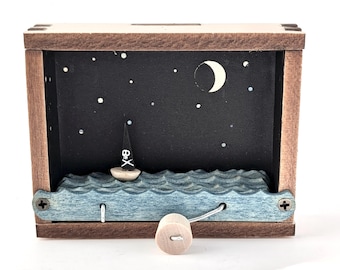 May include: A wooden pirate-themed music box featuring a small sailboat on blue waves under a starry night sky with a crescent moon. The box is rectangular with a crank on the side.