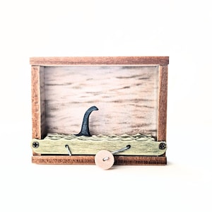 Puede incluir: Una caja de madera con una pequeña figura de criatura marina mítica negra en su interior. La criatura está parcialmente sumergida en una superficie verde ondulada. La caja tiene un pequeño pomo de madera en la parte delantera.