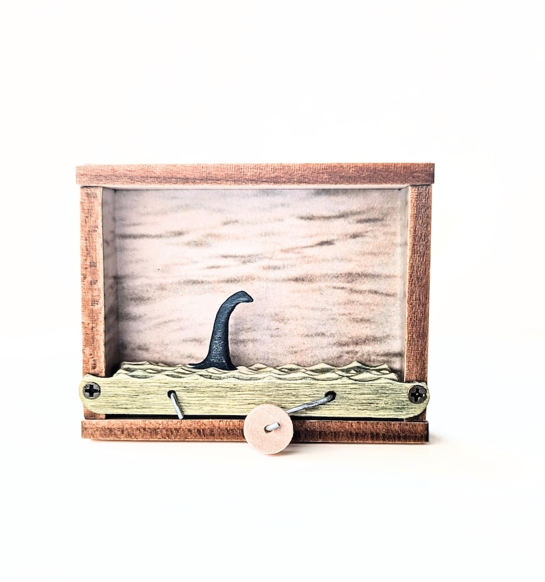 May include: A wooden box with a small, black, mythical sea creature figurine inside. The creature is partially submerged in a green, wave-like surface. The box has a small, wooden knob on the front.