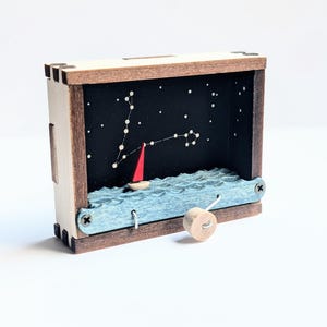 Puede incluir: Caja de sombras de madera con una escena náutica. Un velero rojo navega sobre olas azules bajo un cielo estrellado. La caja tiene un marco de madera clara y un interior oscuro. Un pequeño pomo de madera está unido al lateral.