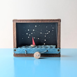 May include: A wooden box featuring a nautical scene with a red-sailed boat on blue waves. The background is a dark blue night sky with a white constellation design. The box has a wooden frame and a small wooden wheel.
