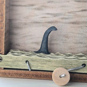 May include: A wooden box with a crank on the side. The box has a cutout in the front that shows a black silhouette of a sea monster's neck and head emerging from the water.