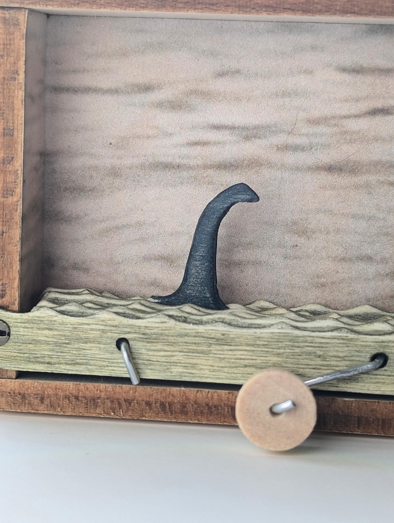 May include: A wooden box with a crank on the side. The box has a cutout in the front that shows a black silhouette of a sea monster's neck and head emerging from the water.
