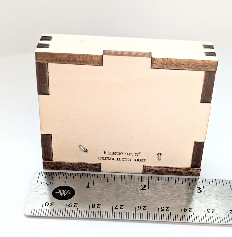 May include: A wooden box with a light brown finish and a dark brown trim. The box is about 2 inches wide and has the text "kinetic art of cartoon monster" printed on the front.
