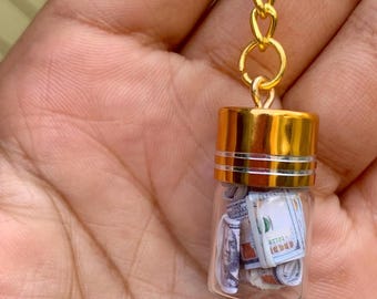 May include: A gold keychain with a small glass jar filled with rolled up US dollar bills. The jar has a gold lid with two silver bands.