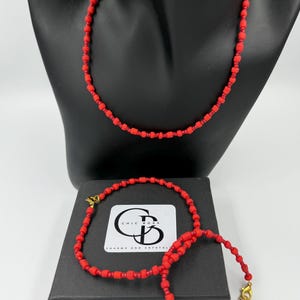 Peut inclure: Un ensemble de trois colliers de perles rouges avec des fermoirs dorés. Les colliers sont présentés sur une boîte noire avec le texte "Chic Boss Charms and Crystals".