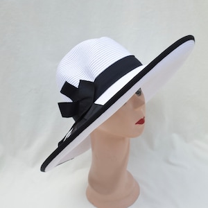 May include: A white and blue striped sun hat with a wide brim. The hat features a black ribbon band and a large black bow. The brim is trimmed with black. The hat is displayed on a mannequin head.